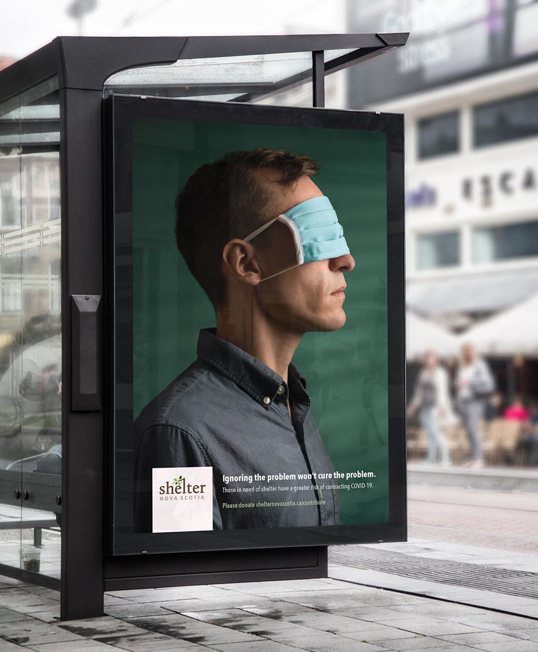 Bus stop advertisement on busy street for Shelter Nova Scotia with man blinded by COVID face mask resting over his eyes. 