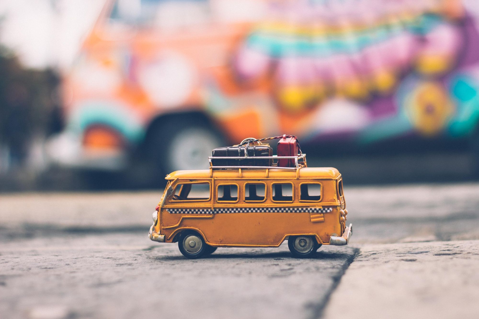 Miniature yellow Volkswagen with luggage strapped onto roof sitting on pavement with blurred life-size Volkswaegen behind